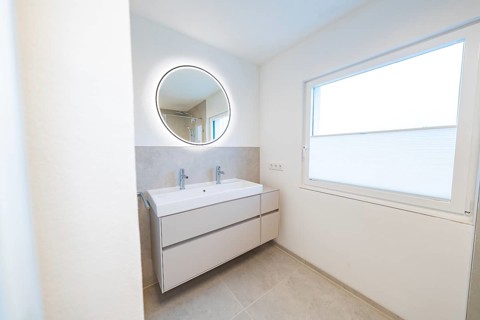 Bright bathroom with round illuminated mirror and double washbasin in front of subtle shades of
