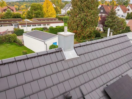 Roof with anthracite-colored tiles and modern chimney, behind it quiet residential area with autumn