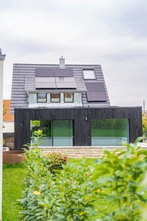 Front view of a house with solar panels and black extension with glass front, well-kept garden and