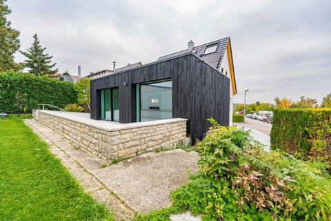 Black, modernly clad extension with large glass areas on lawn and stone wall in a quiet suburban