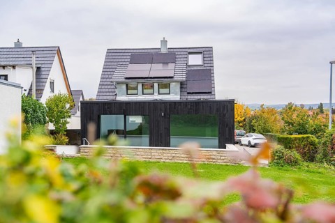 House with solar panels and black extension, view over garden to quiet street with parked car,