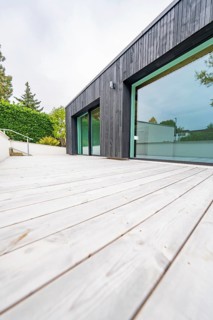 Deep view along the wide wooden terrace of the modern, black-clad building with floor-to-ceiling