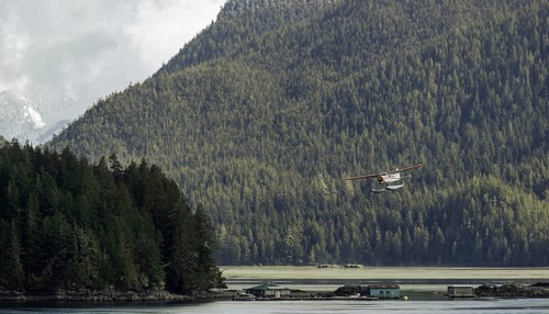 A seaplane flies over a tranquil lake surrounded by dense forest and mountains under an overcast