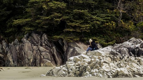 A person sits on rocky coastline surrounded by trees under a cloudy sky in Tofino, British