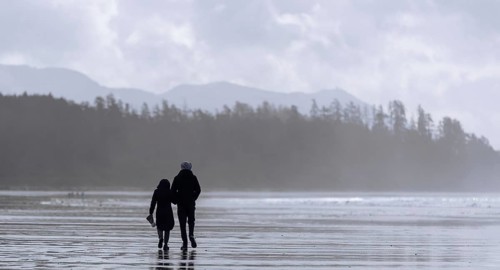 A couple strolls on a misty beach with forest and mountains in the distance in Tofino, British
