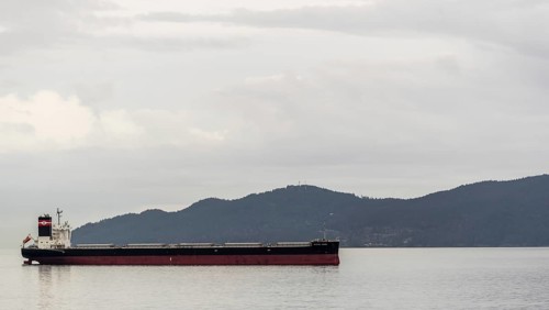 A solitary cargo ship sails by a mountainous backdrop under cloudy skies in Vancouver, British
