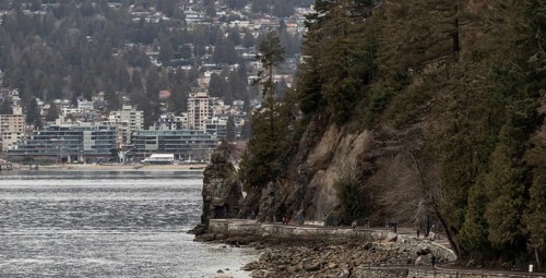 Rocks and trees line a coastal path in Stanley Park with urban buildings in the distance in