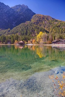 Emerald green, clear shore water with visible stones, colorful autumn trees and a boathouse are