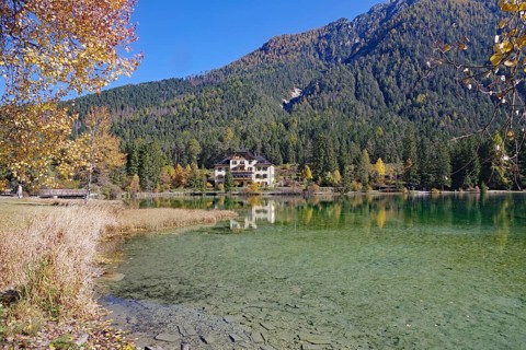 Wide shore with shallow, crystal-clear water, opposite is a villa in the sunshine in autumn forest,