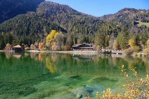 Clear mountain lake with emerald green water, autumnal forest and hut on the shore under blue sky,