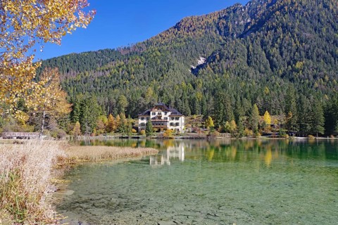Clear shore water of an alpine lake, opposite is a villa reflected against colorful autumn forests