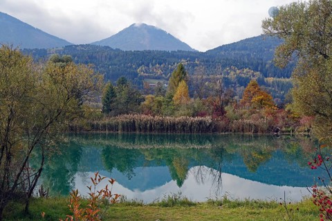 Quiet lake with turquoise-green water, autumnal shore and mountain scenery under cloudy sky, Drau,