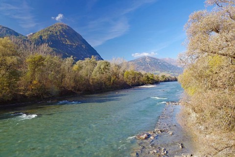Clear river with gravel bank between autumn trees, mountains in the background under blue sky,