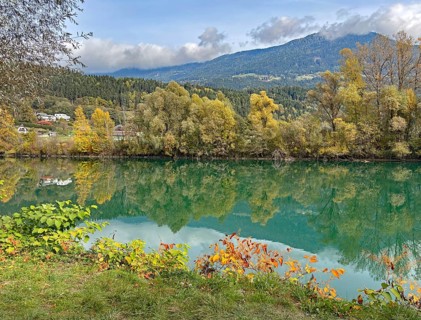 Calm river reflecting autumnal trees and mountains under cloudy sky, Drau, Drauradweg, Carinthia,