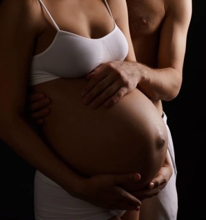 Pregnant young long haired woman staying close to her husband both tenderly looking at her belly