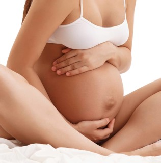 Young long haired pregnant brunette in white top sitting on bed posing holding her hands on her