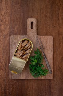 An open can of sprats, appetizer, on a wooden cutting board, with a fork and herbs, top view