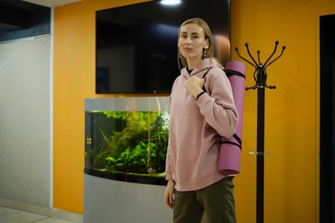 A woman in a pink hoodie stands in a fitness studio, holding a yoga mat. An aquarium with aquatic