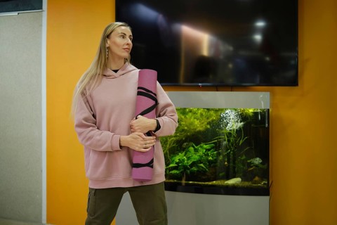 A woman stands in a fitness studio, holding a yoga mat, looking inspired. An aquarium filled with
