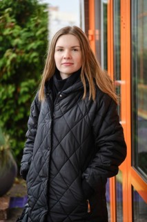A young woman wearing a black quilted coat stands confidently outside a building. She has long hair