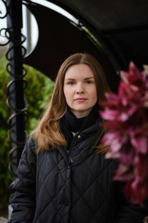 A young woman with long hair poses confidently near vibrant flowers in a garden. The scene captures