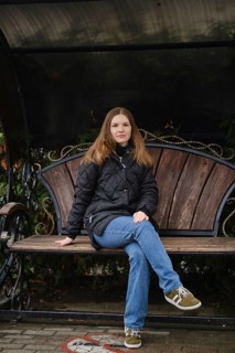 A young woman sits calmly on a wooden bench in a park surrounded by autumn foliage. She wears a
