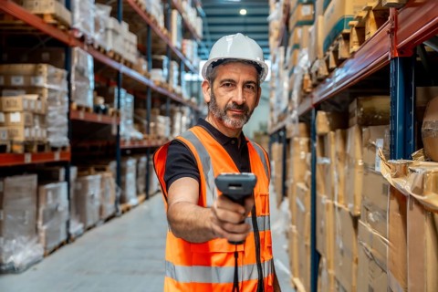 Warehouse worker wearing safety helmet and vest, holding scanner, checking inventory and managing