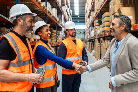 Business executive is shaking hands with a diverse team of warehouse workers wearing safety vests