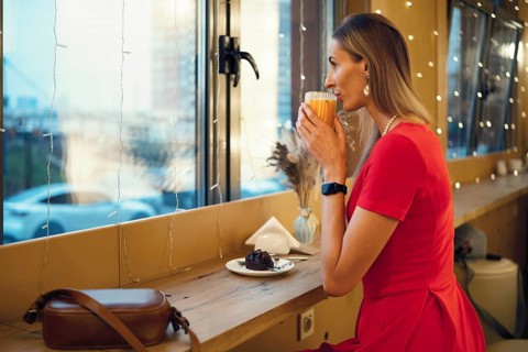 A woman in a red dress sits by the window of a cafe, sipping a warm drink and enjoying a dessert.