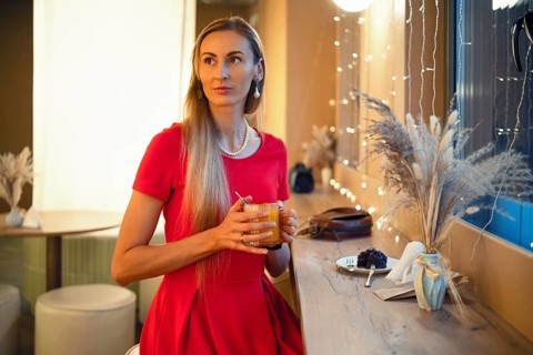 A woman with long hair sits at a bar in a cozy cafe, holding an orange drink. She looks thoughtful