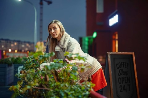 A woman leaning on the railing decorated with the plants outside a coffee shop in the evening. The
