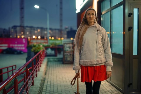 A woman dressed in a light jacket and red skirt walks along a sidewalk near shops in a city. The