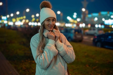 A woman stands outside on a city street during the evening, wearing a warm jacket and a hat with a