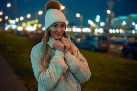 A woman wearing a cozy coat and hat stands outside on a cool evening. The city lights twinkle in