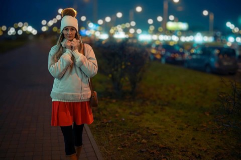 A woman walks on a city sidewalk at night, wearing a warm white jacket and a beanie. The street is