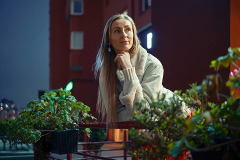 A woman with long hair leans on a railing surrounded by lush plants, reflecting on a lively city
