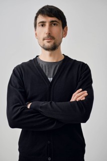 A man stands with arms crossed, portraying confidence. He wears a black cardigan over a gray shirt.