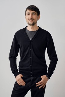 A man stands confidently with his hands on his hips, wearing a black cardigan over a t-shirt. The
