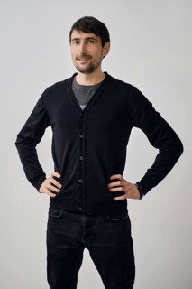 A man poses confidently with his hands on his hips, wearing a black cardigan over a gray shirt. He