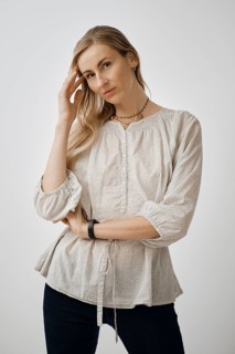 A pensive woman is standing with one hand touching her hair while wearing a light, textured blouse.