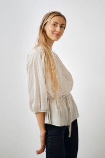 A blonde woman model stands playfully in a light-colored blouse with bell sleeves. She wears dark