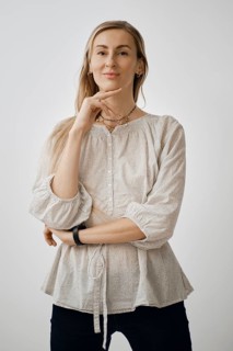 A woman stands in a relaxed pose with one hand on her chin, wearing a light-colored, casual blouse.