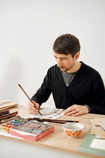 A male artist focuses on his work at a wooden table filled with various art supplies. He uses