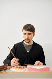 A focused artist is working diligently on a paper in a clean, bright studio. He uses a brush to add