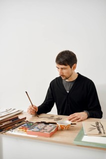 A focused artist sits at a wooden table, working on an intricate drawing with a brush