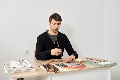 An artist sits at a wooden table, focused on painting with watercolors. The studio is bright and