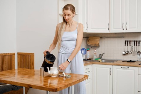 A woman stands in a modern kitchen pouring hot water from a kettle into a teapot. She wears a light