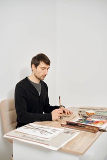 A person is focused on drawing at a tidy desk filled with art supplies and books. The bright room