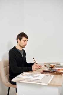 In a light-filled home studio, a man sits at a desk focused on drawing. He uses a brush to create