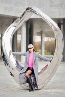 A stylish woman poses sitting on metallic sculpture in a city, showcasing urban street fashion